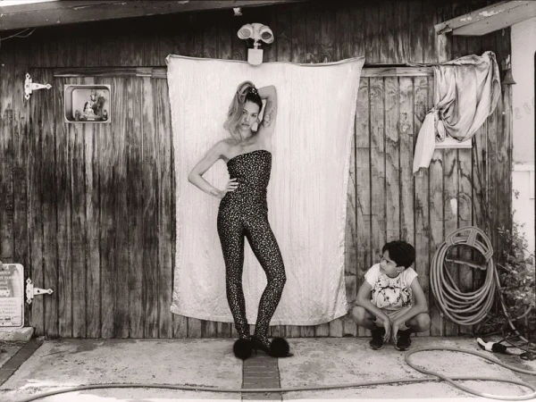 Black-and-white image of a woman in a patterned jumpsuit and a child squatting in front of a wooden wall, garden hose, and a white sheet.