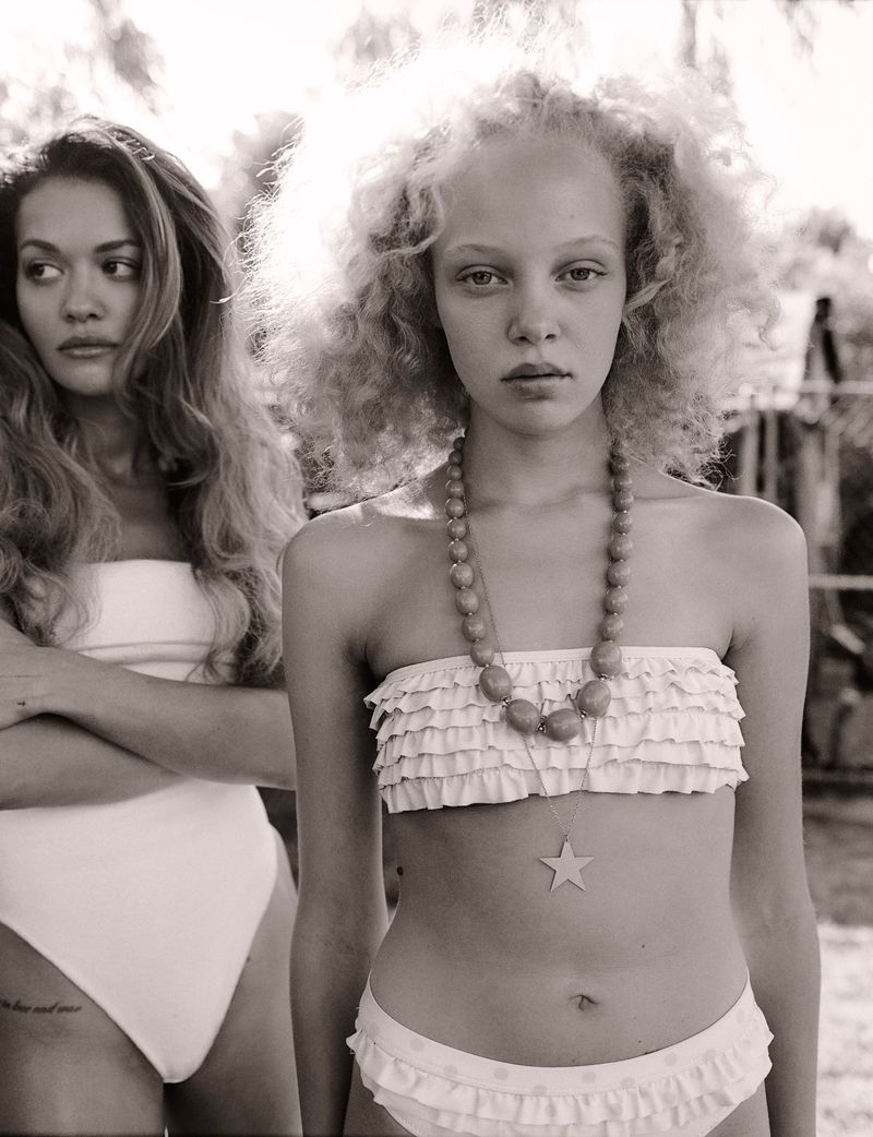 Two women in swimwear pose outdoors in a black and white fashion portrait, one with curly blonde hair and wearing a beaded necklace.