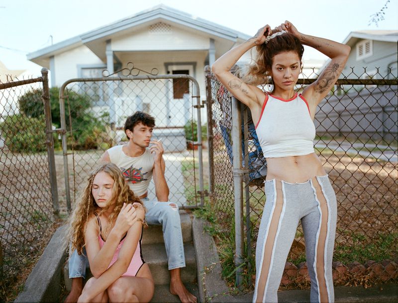 Three young adults pose outside a house with a chain-link fence; one woman stands while the other woman and man sit on the steps.