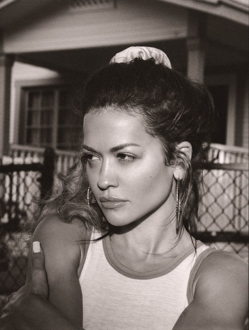 Black and white portrait of a woman wearing a tank top, hoop earrings, and a hair scrunchie, posing in front of a house.