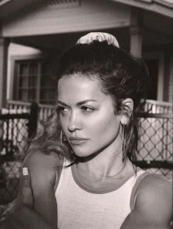 Black and white portrait of a woman wearing a tank top, hoop earrings, and a hair scrunchie, posing in front of a house.