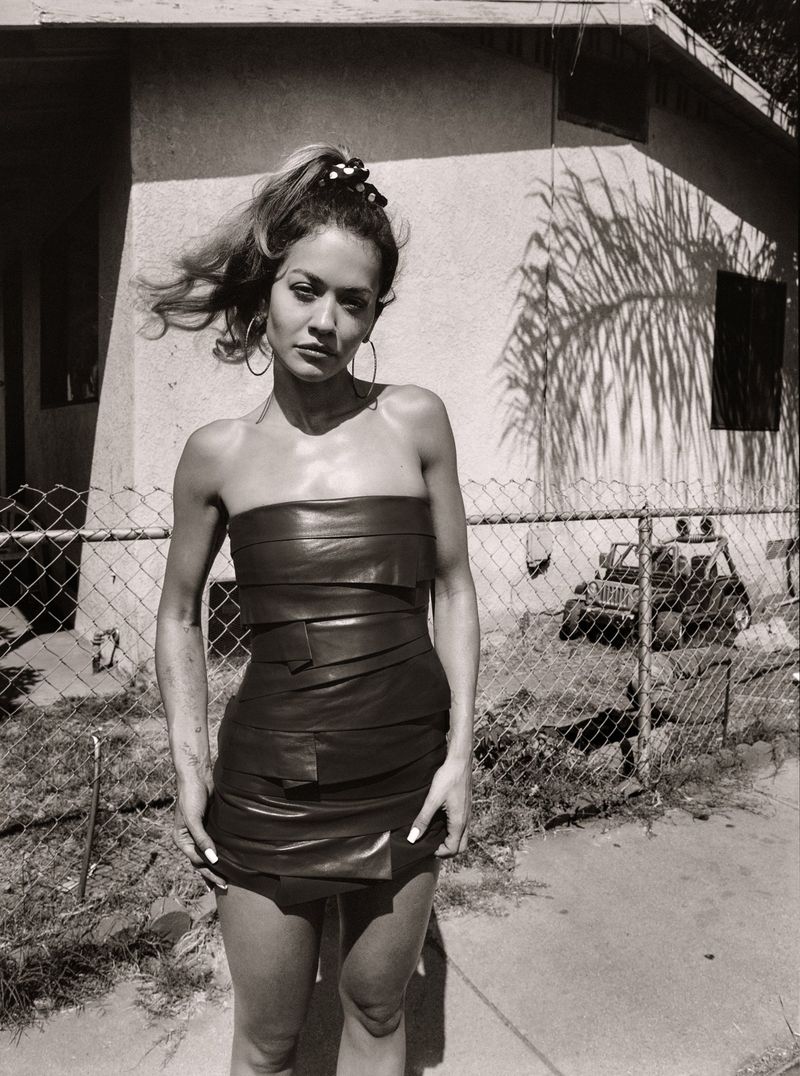 Woman in a black leather dress posing outdoors in front of a house with a chain-link fence, captured in a monochrome outdoor portrait.