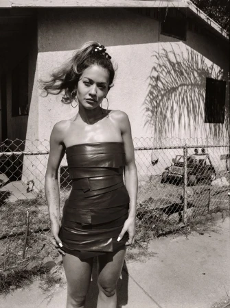 Woman in a black leather dress posing outdoors in front of a house with a chain-link fence, captured in a monochrome outdoor portrait.
