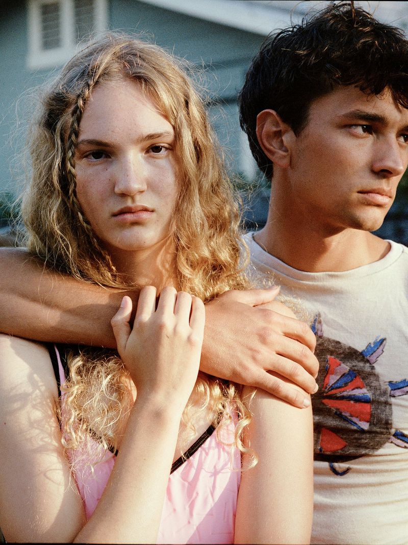 A young adult couple poses outdoors with arms around each other. The woman has blonde hair and a pink tank top; the man wears a graphic t-shirt.