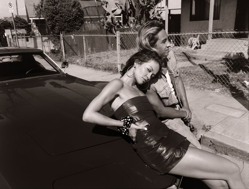 Black and white portrait of a woman in a strapless dress reclining on a vintage car with a man in a denim vest standing behind her.