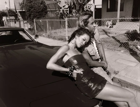 Black and white portrait of a woman in a strapless dress reclining on a vintage car with a man in a denim vest standing behind her.