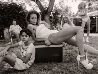 Rita Ora posing outdoors in a white strapless swimsuit and heels seated on a cooler box with several other people in the background