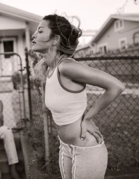 Rita Ora wearing a white tank top and grey trousers posing outdoors by a chain-link fence in a black and white portrait.