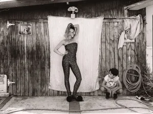 Rita Ora posing in a patterned catsuit before a makeshift fabric backdrop with a seated young boy nearby