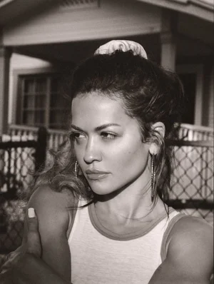 Monochrome portrait of Rita Ora with hair up, wearing hoop earrings and a tank top against an outdoor residential building background