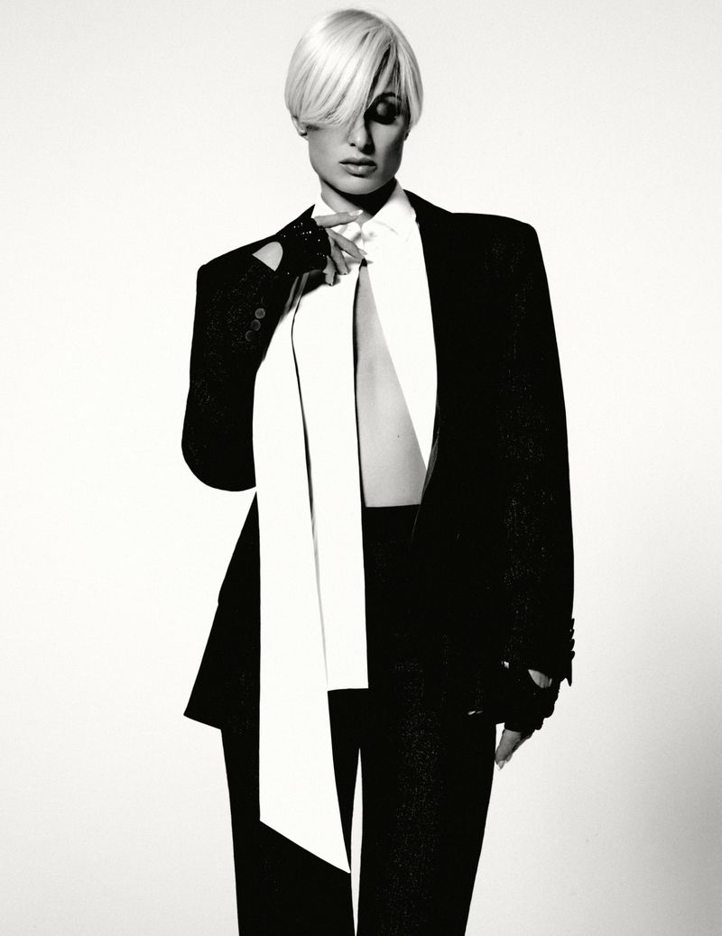 Monochrome portrait of a blonde woman wearing a black suit and white tie, posing in a photo studio against a white backdrop.