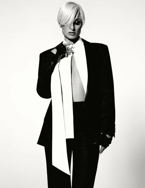 Monochrome portrait of a blonde woman wearing a black suit and white tie, posing in a photo studio against a white backdrop.
