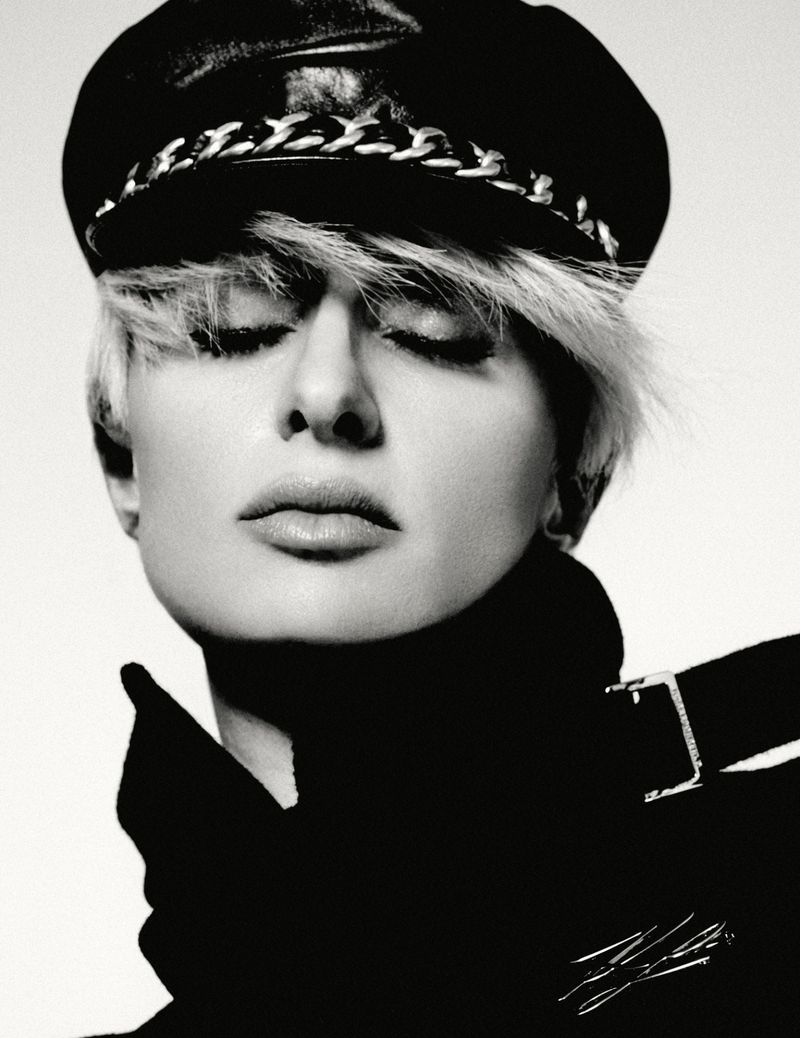 Black and white portrait of a blonde adult female with closed eyes wearing a black leather hat and coat against a white background.
