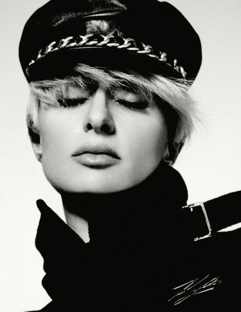 Black and white portrait of a blonde adult female with closed eyes wearing a black leather hat and coat against a white background.