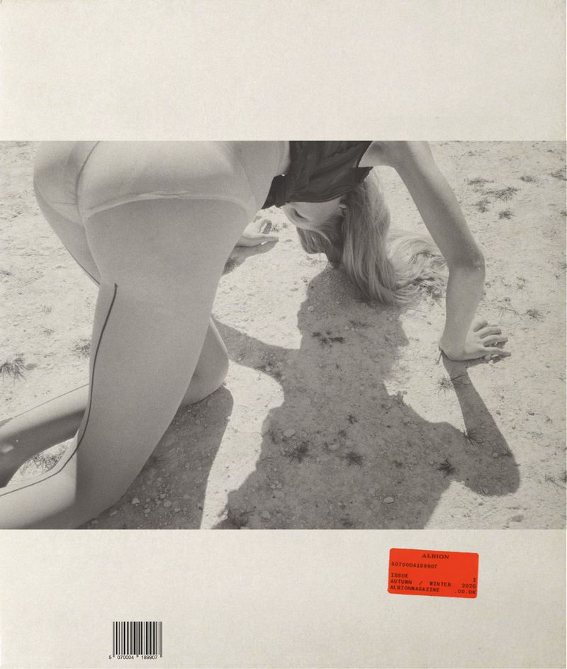 Monochrome photograph of a blond woman kneeling on sand, showcasing her back and posterior, with her shadow cast on the ground.