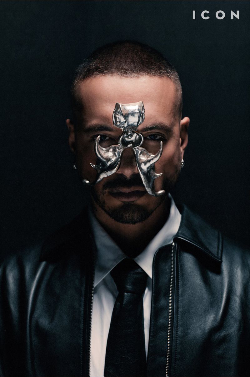 Portrait of a man wearing a silver mask-like face accessory, white shirt and tie, and a black leather jacket against a dark background.
