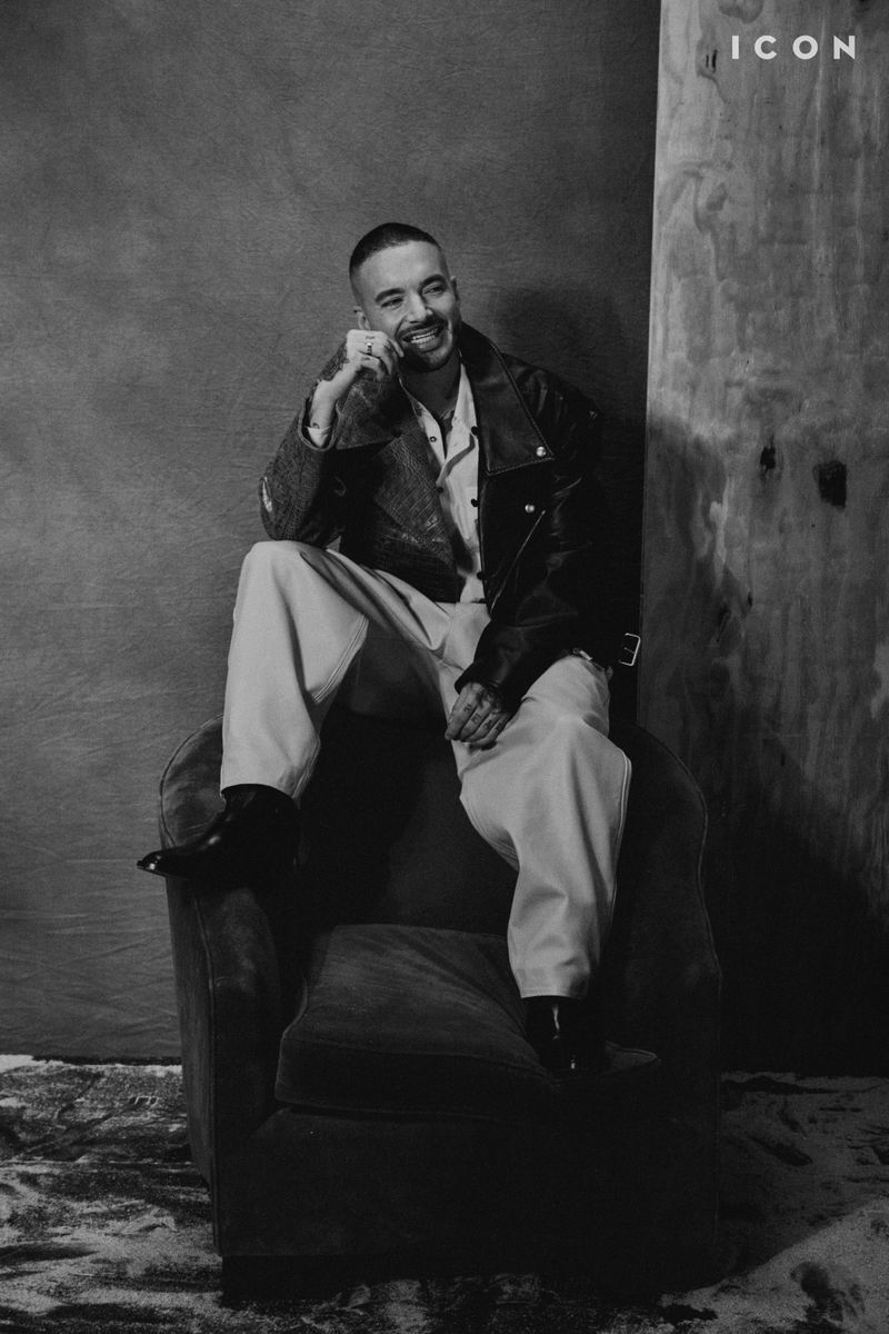 Black and white portrait of a smiling man in a jacket, shirt and pants sitting in an armchair in front of a textured wall.