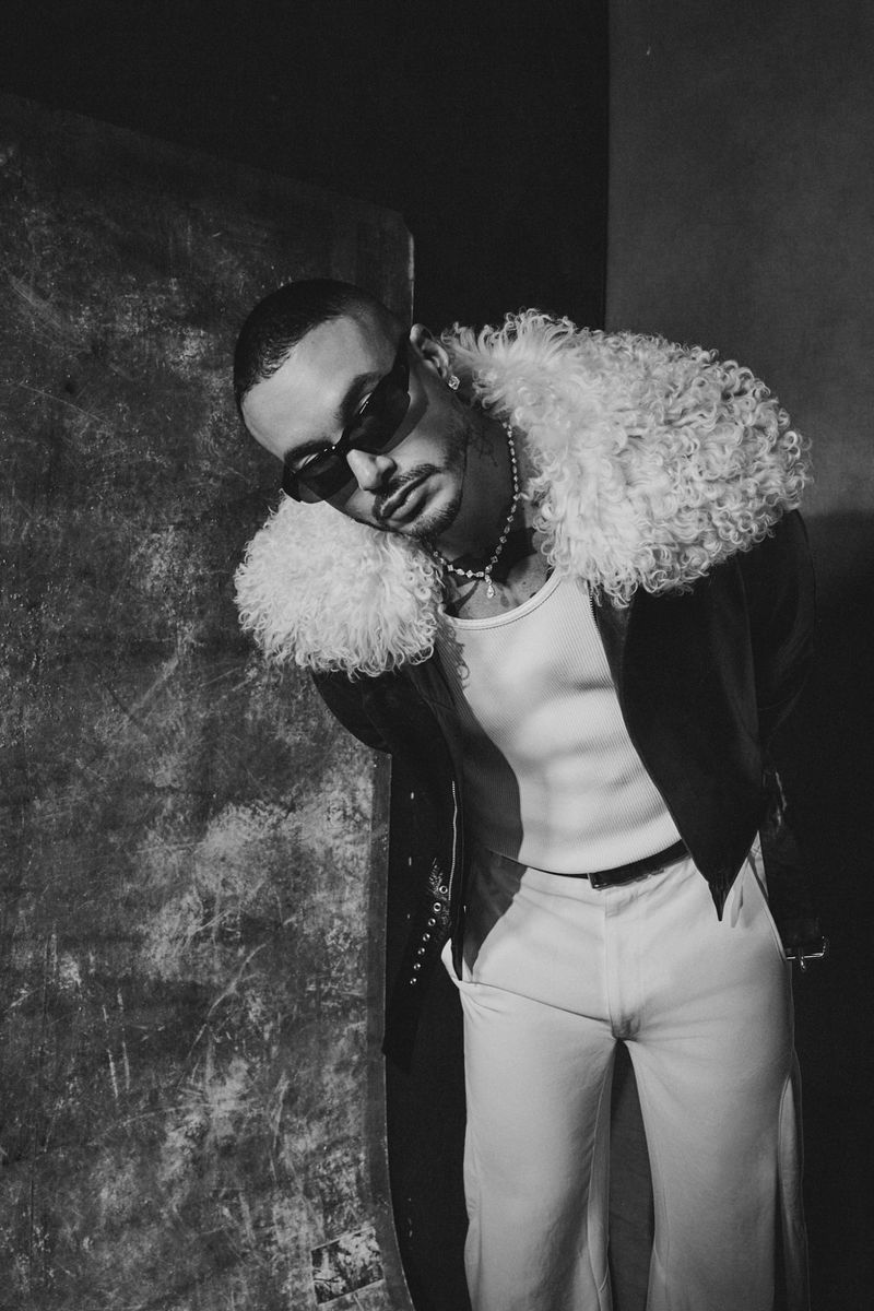 Monochrome portrait of a man in sunglasses wearing a shearling-collared coat leaning against a textured wall in a studio setting.