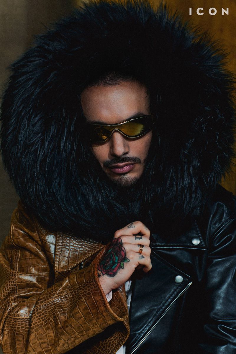 Fashionable adult male portrait. He wears sunglasses, a fur-trimmed hood, and a two-tone jacket. Rose tattoo is visible on his hand.