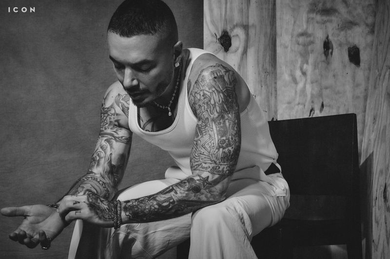 Monochrome portrait of an adult male with tattoos in a white tank top, sitting on a chair against a textured background.