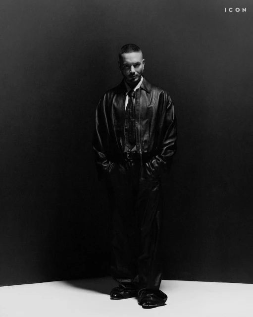 Monochrome studio shot of a man in a leather jacket and pants, standing against a gradient dark backdrop with a white floor.
