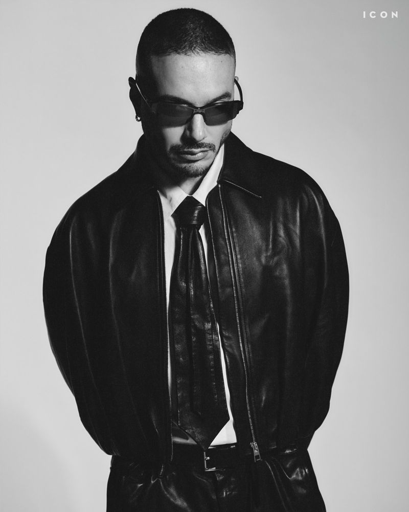 Monochrome portrait of an adult male with sunglasses, leather jacket, and necktie in a studio setting against a neutral backdrop.