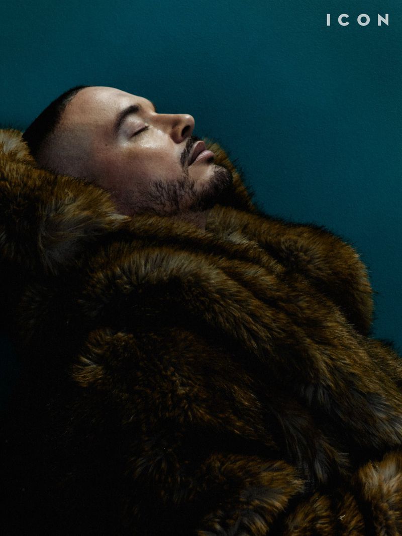 Portrait of a man with short hair and a beard, wearing a fur coat, lying against a dark teal background. The image has a magazine cover feel.