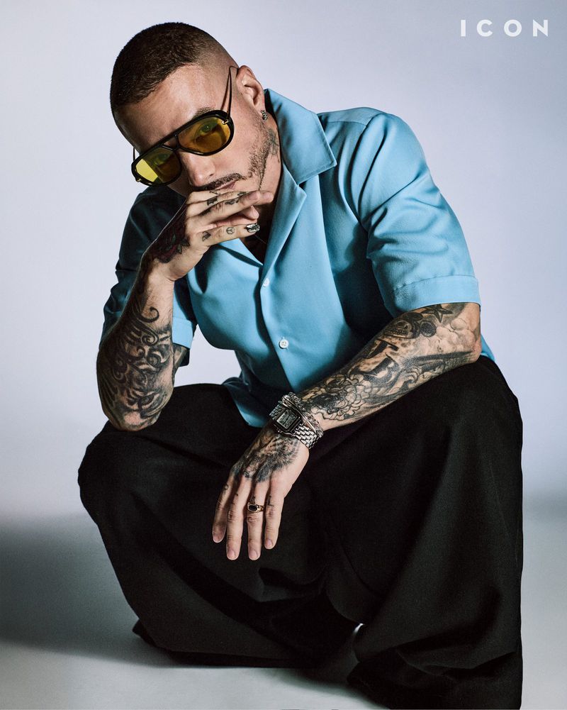 Stylish man with tattoos and yellow sunglasses crouching in a studio, wearing a light-blue button-down shirt and black pants.