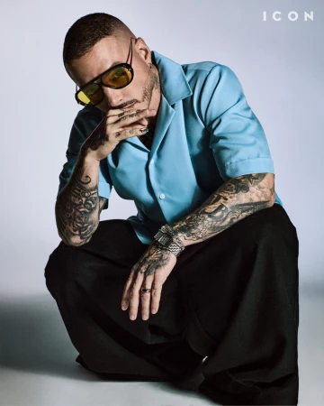 Stylish man with tattoos and yellow sunglasses crouching in a studio, wearing a light-blue button-down shirt and black pants.