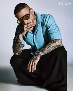 J Balvin in a light blue short-sleeve shirt and black trousers wearing tinted sunglasses against a grey studio backdrop