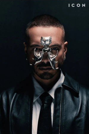 Portrait of J Balvin wearing a sculptural silver face piece, white shirt, black tie, and leather jacket against a dark background