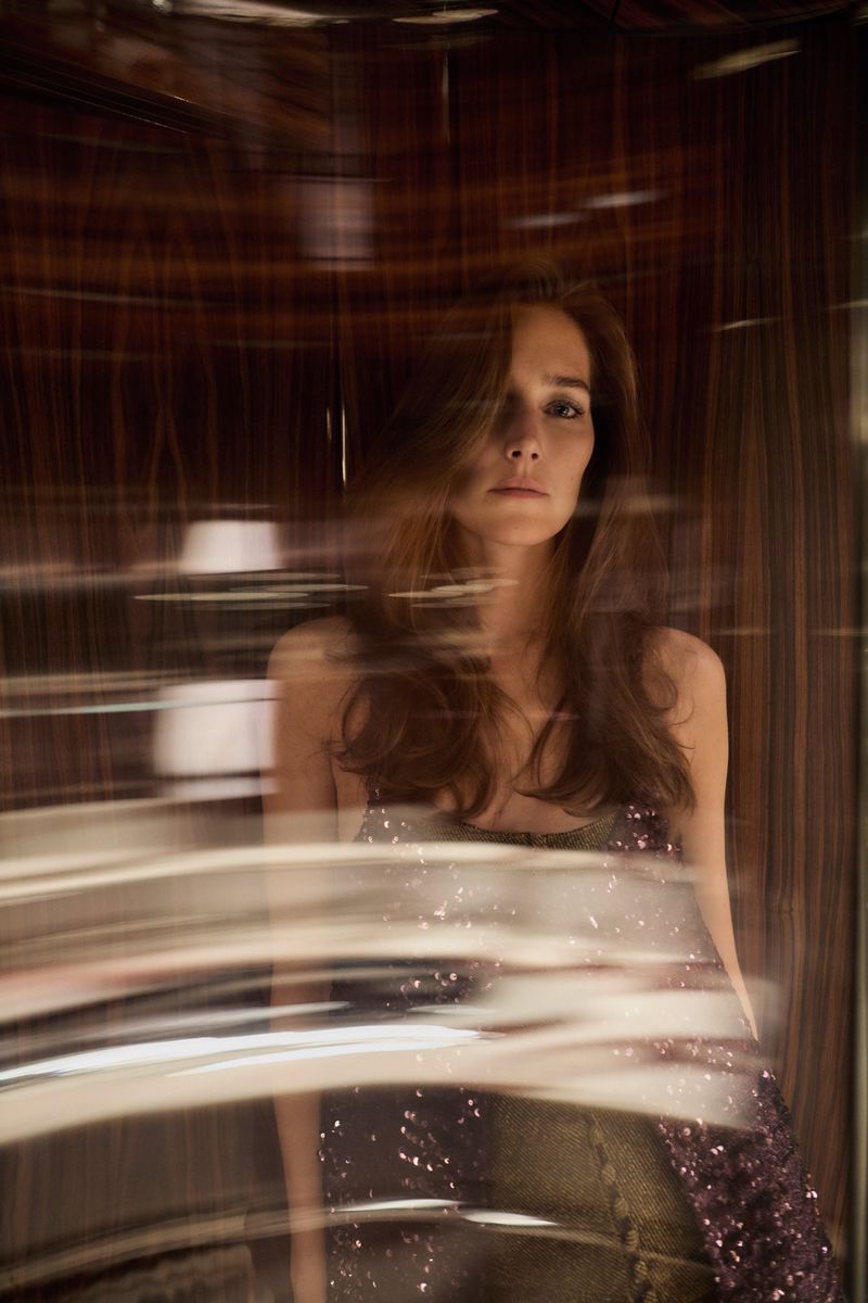 Woman with long brown hair and sequin dress poses in an interior setting with blurred reflective surfaces and warm lighting. 