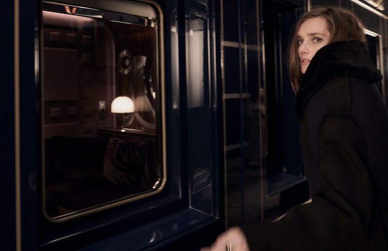 Woman with brown hair in dark coat stands beside a train car with a lit interior visible through the window, in dim lighting.