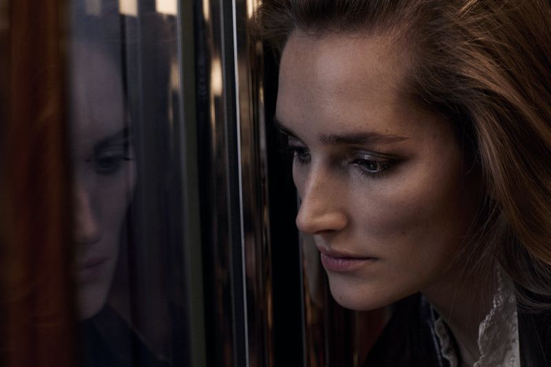 A close-up portrait showcases an adult female with long brown hair and a thoughtful expression, reflecting in a pane of glass.