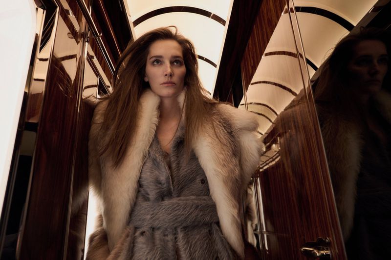 Woman with long hair wearing a fur coat stands in a reflective corridor with wood paneling and curved lighting above.