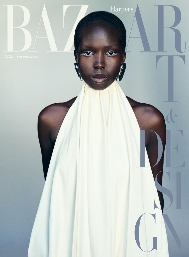 Fashion editorial featuring a dark-skinned model with white eye makeup wearing a cream-colored draped garment against a light gray backdrop.