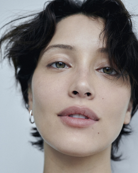 Close-up portrait of a woman with short dark hair and natural makeup against a plain grey background