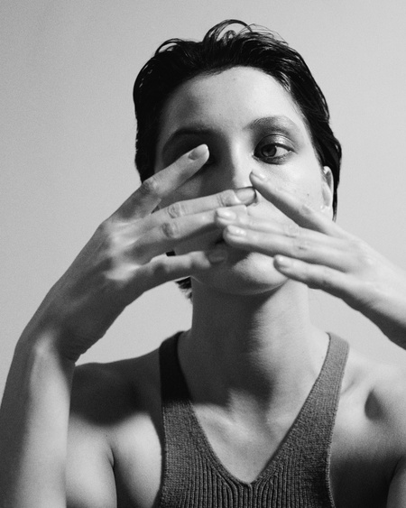 Close-up black and white portrait of a model with slicked-back hair holding hands near face, wearing a tank top against a plain background.
