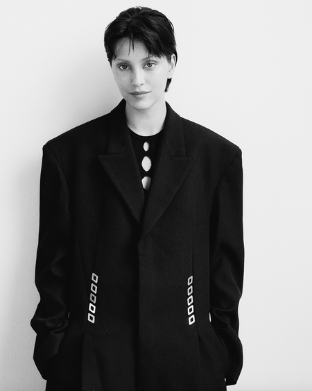 A woman with short dark hair wearing a large black coat poses in front of a white background for a fashion-style portrait.