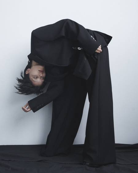 A model in a black suit strikes a dynamic pose in a studio. The dark clothing contrasts with the clean, light backdrop, creating a strong visual.