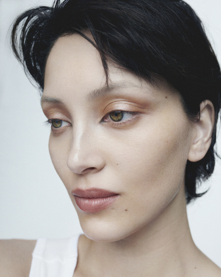 Close-up portrait of a woman with short dark hair and makeup, wearing a white top against a neutral background, focusing on beauty and skincare.