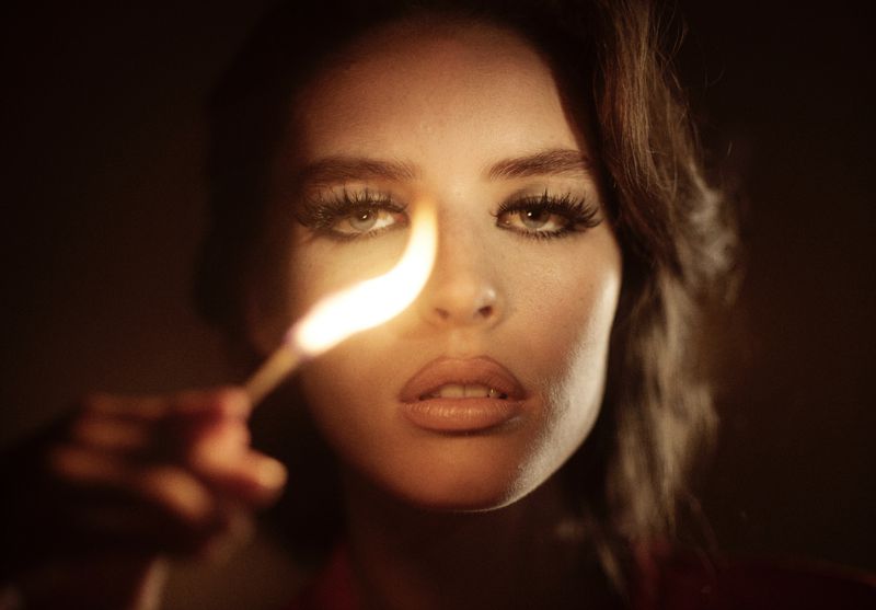 Close-up portrait of a woman holding a lit match, casting warm light on her face, with a dark background creating a dramatic effect.