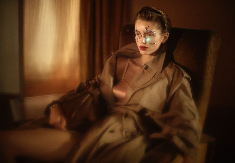 Adult female with glitter makeup posing in a trench coat, sitting in an armchair by a window, illuminated with golden light in a studio.