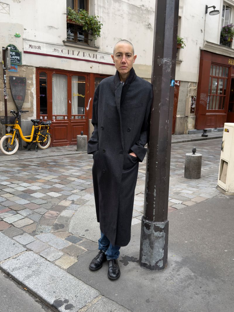 Adult female stands in a long dark coat and jeans on a cobblestone street in Paris next to a metal pole, yellow bicycle behind her.