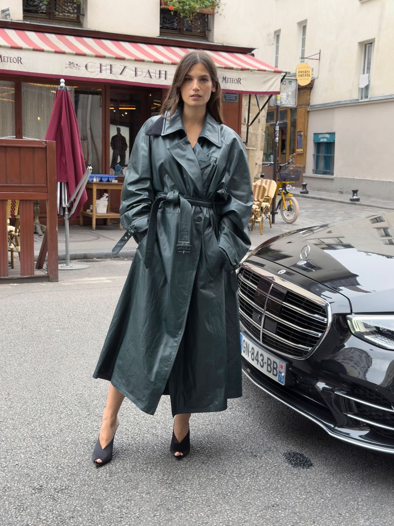 Woman in a shiny green trench coat standing next to a black car on a city street with an awning and building in the background.