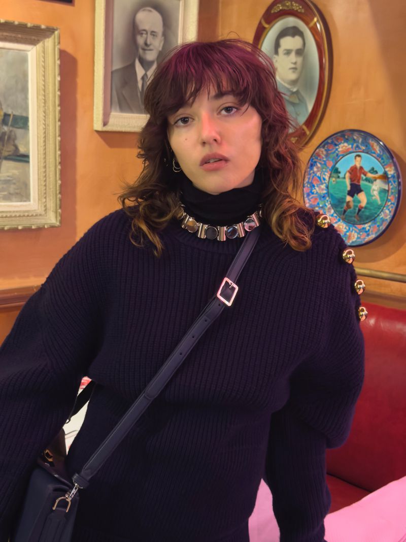 Portrait of a woman with bangs wearing a black turtleneck sweater and necklace with gold accents, in front of framed pictures.