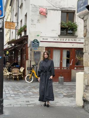 A woman wearing a long dark belted coat standing on a cobblestone street in front of a Parisian restaurant storefront.