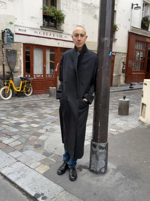 Person with a shaved head wearing a long dark grey coat leaning against a post on a cobblestone European street.
