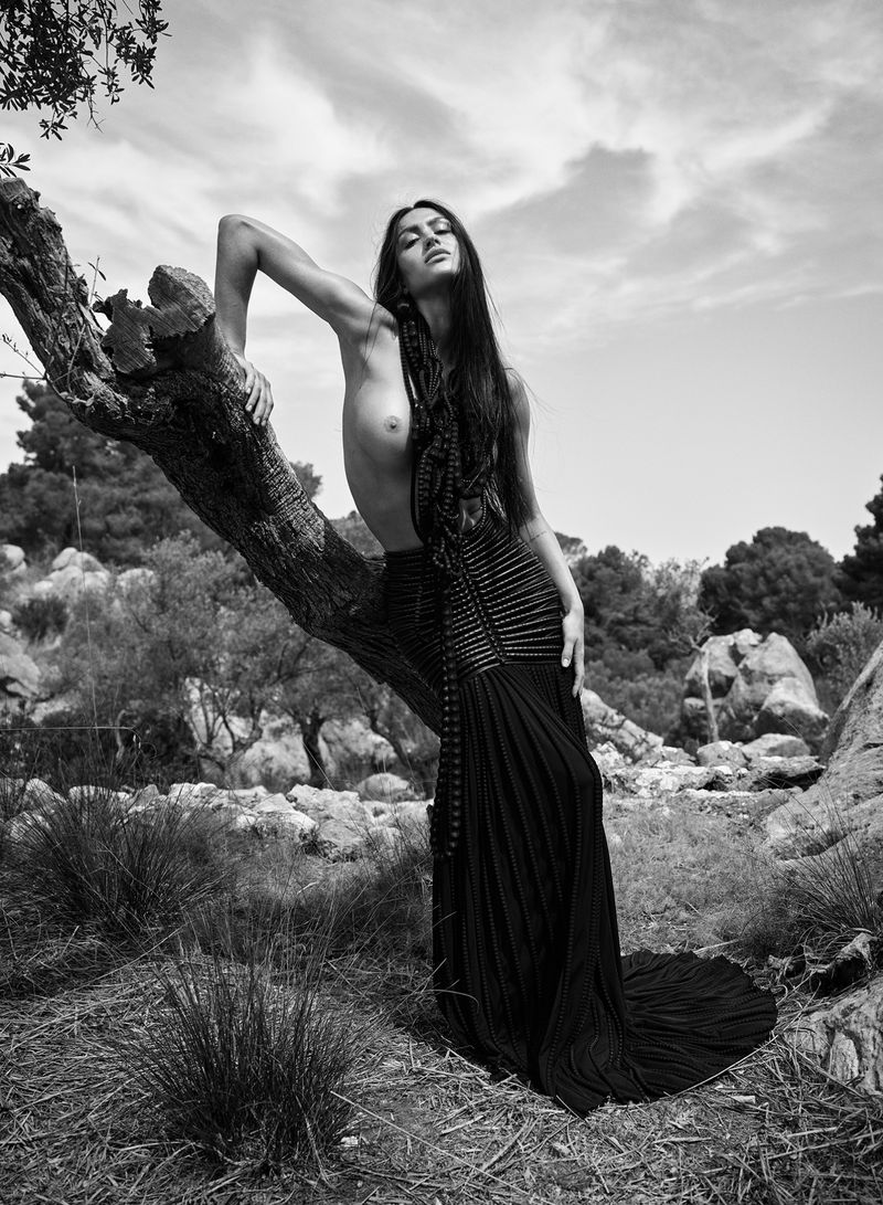 Topless woman in a flowing black dress reclines on a tree branch outdoors against a backdrop of lush greenery and cloudy sky in black and white.
