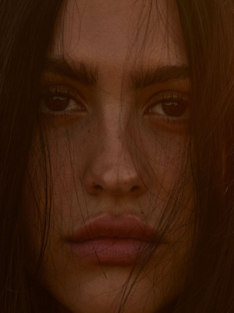 Close-up portrait of a young adult female with long, dark hair partially obscuring her face, brown eyes, and full lips in warm, soft lighting.